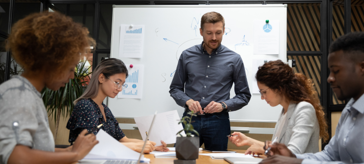 man standing in front of group of business people, looking over documents and auditing data