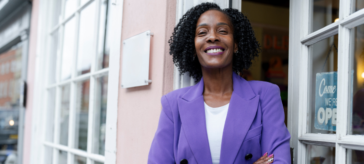 black woman standing outside business she owns with purple jacket on smiling with arms folded